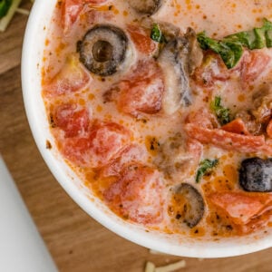 close up of pizza soup with sausage pepperoni olives in a white bowl on a wooden cutting board