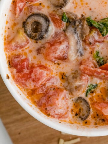 close up of pizza soup with sausage pepperoni olives in a white bowl on a wooden cutting board