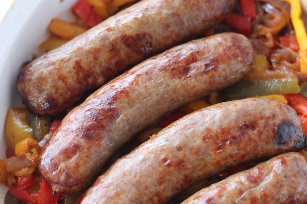 how to cook bratwurst without a grill, brats in a white dish on a bed of cooked peppers