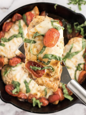 the baked caprese chicken in a skillet with one chicken breast topped with balsamic and basil on a spatula.