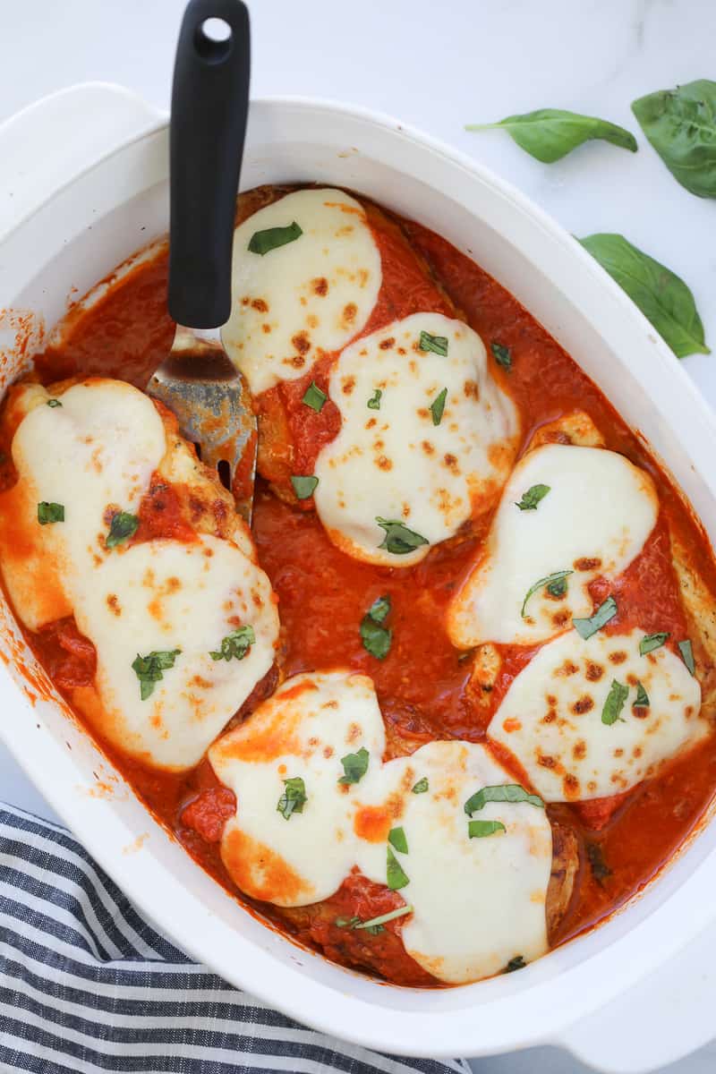 making chicken parmesan no breading is healthier, shown here a a vertical photo in a white pan on a white background