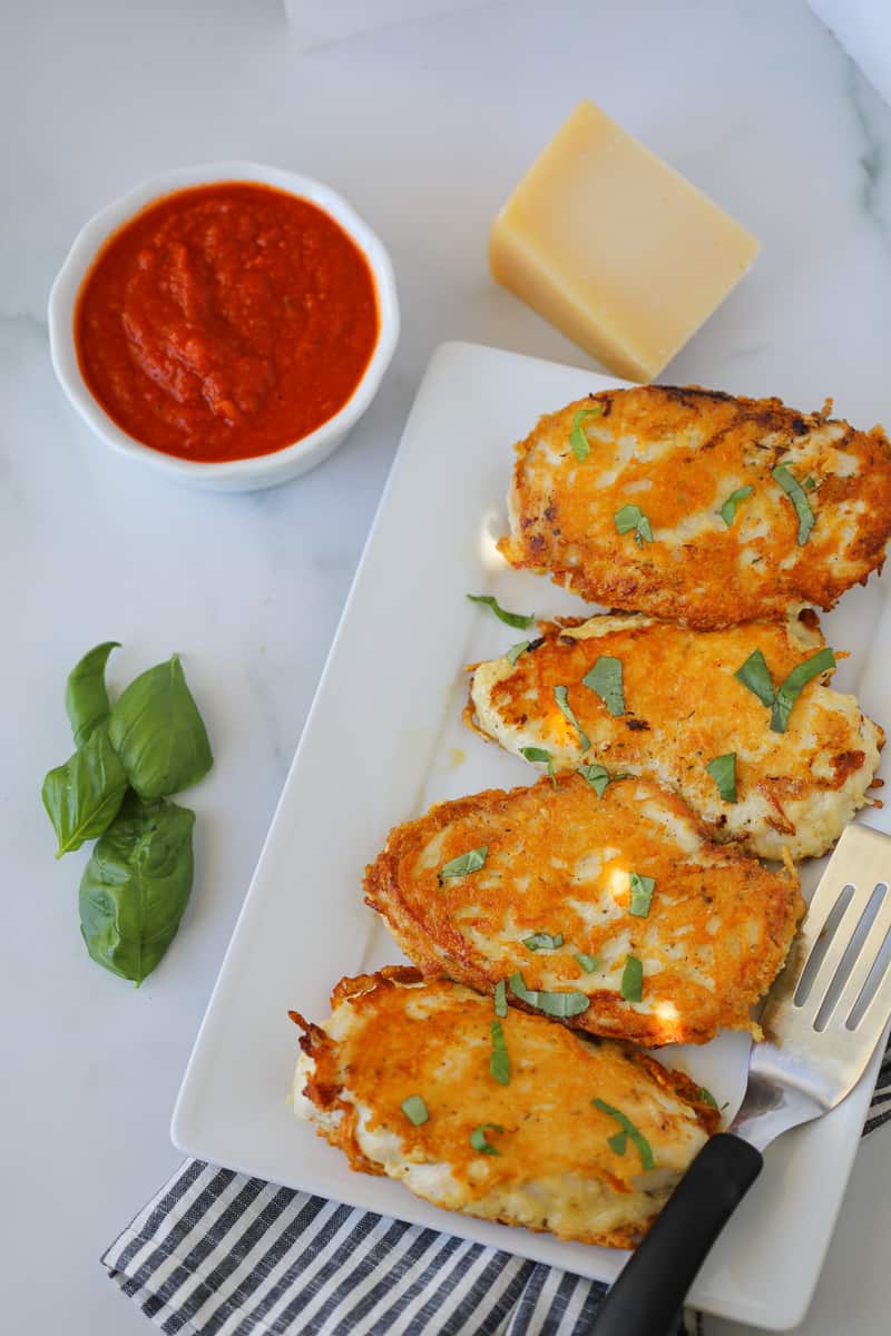 low carb parmesan crusted chicken after cooking on a white platter with sauce, basil, and cheese on the side