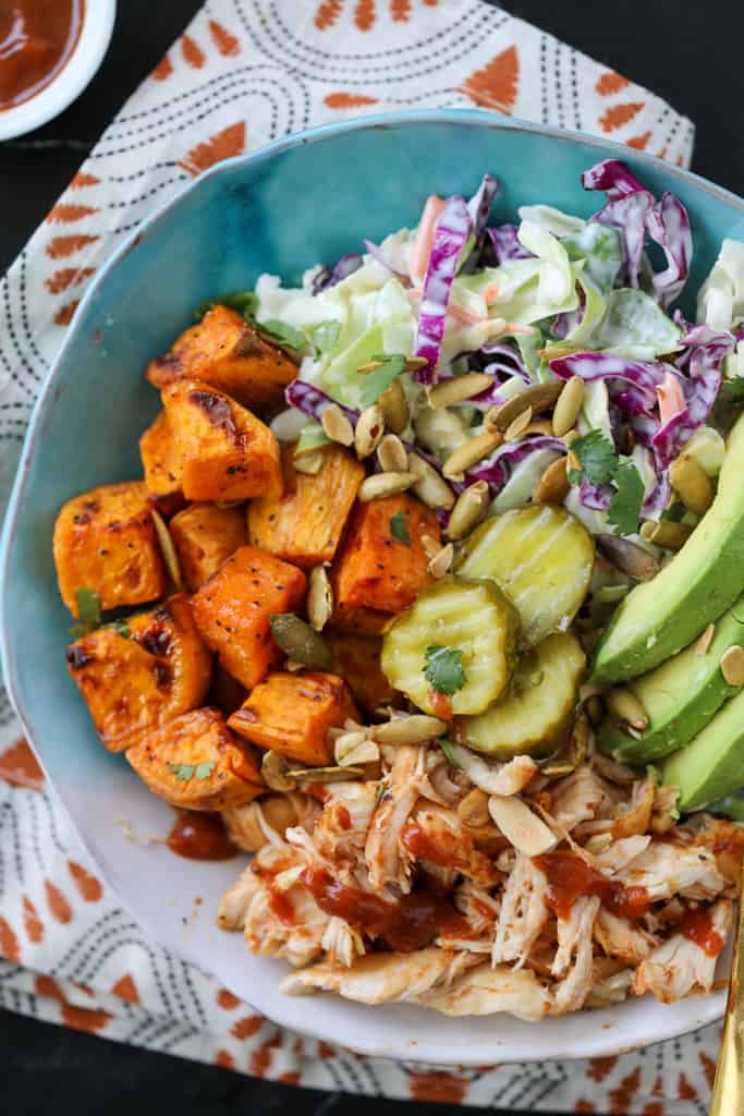 bbq chicken sweet potato bowl in a blue bowl with garnishes of pickles coleslaw and avocado