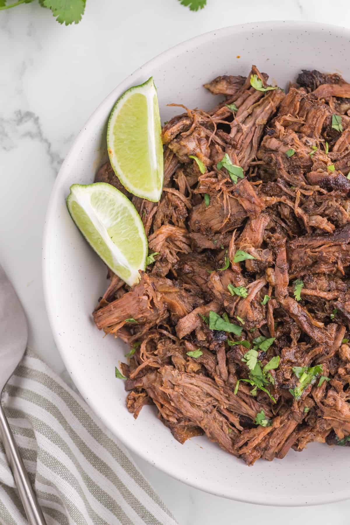 A bowl of fresh made barbacoa beef with a side of lime wedges.