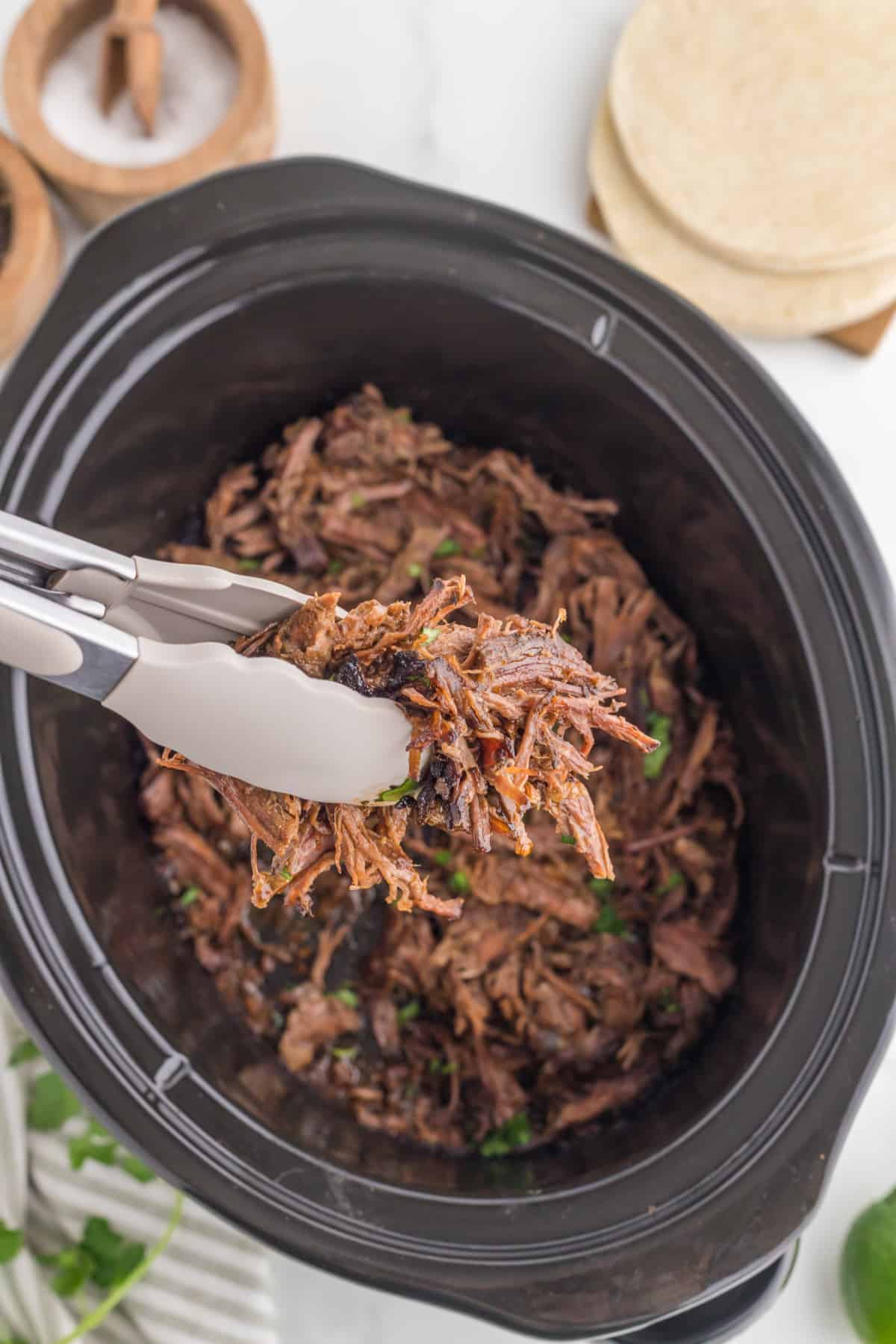 barbacoa beef shredded in a crockpot with tongs pulling it out.