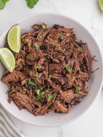 A bowl of fresh made barbacoa beef with a side of lime wedges.