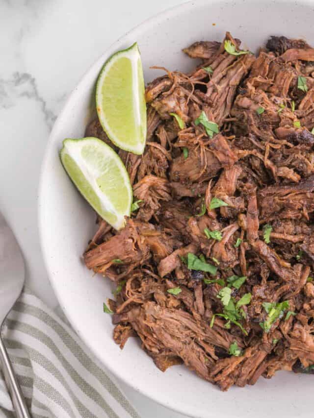 A bowl of fresh made barbacoa beef with a side of lime wedges.