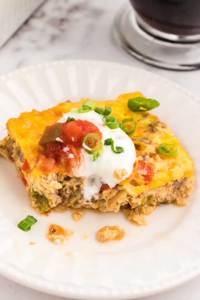 A square slice of keto breakfast casserole on a white plate topped with sour cream and salsa.