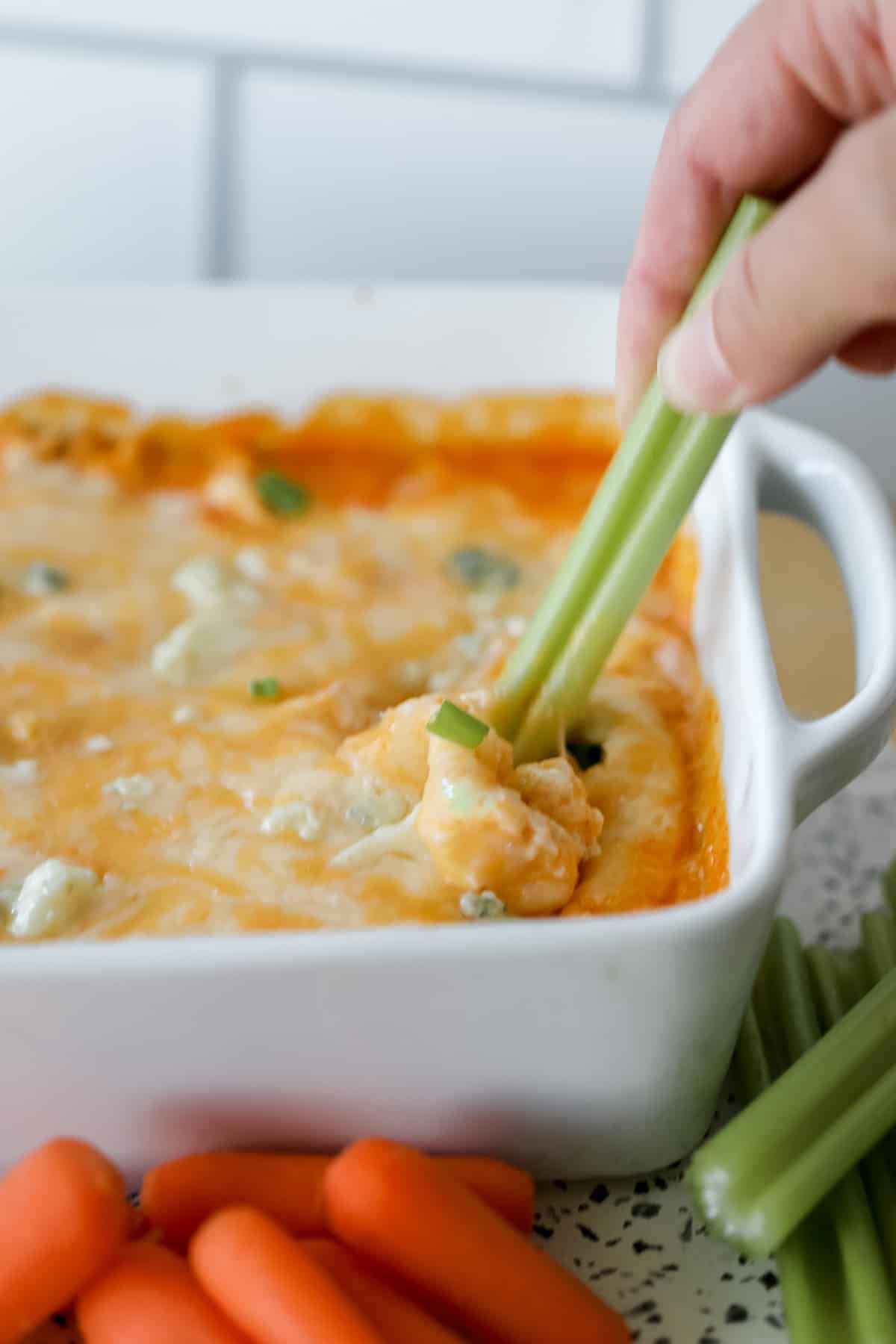 healthy buffalo chicken dip on a white cutting board with celery getting dipped into the dip with a hand