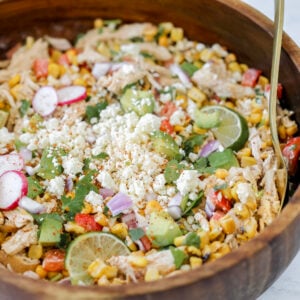 the mexican corn and chicken salad in a wooden bowl topped with cotija cheese lime cilantro and tajin